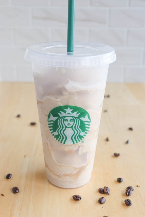 A tall plastic cup filled with iced coffee sits on a light wooden table. The cup has the Starbucks logo and a green straw. Coffee beans are scattered around the table. The background features a white tiled wall.