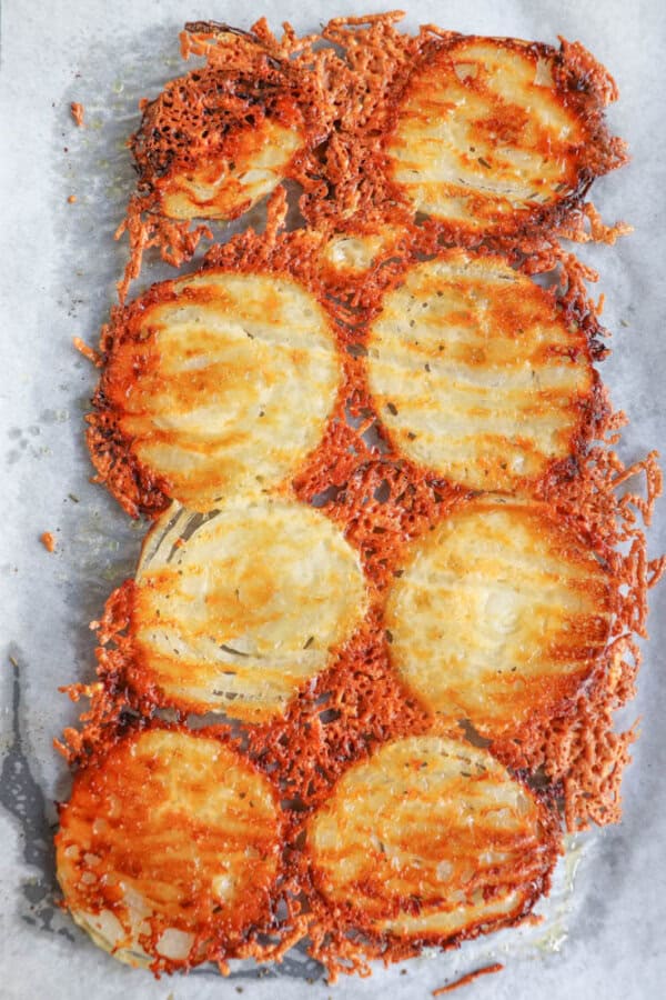 Golden-brown roasted slices of potato with crispy edges are arranged on a baking tray lined with parchment paper. The potato slices are seasoned and topped with melted crispy cheese.