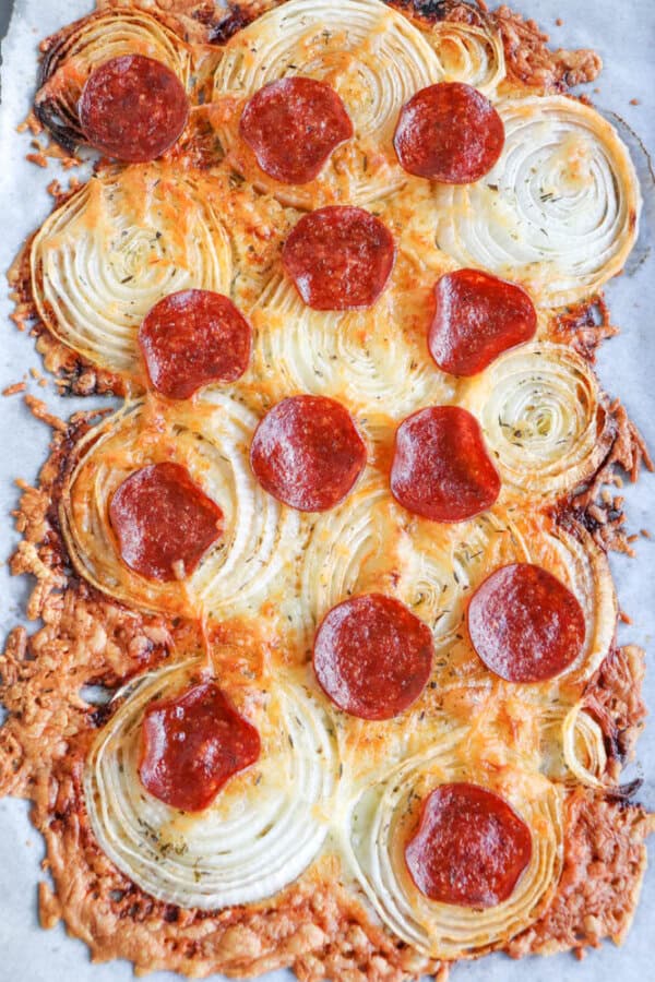 A pizza-like dish topped with melted cheese, crunchy onion ring chips, and pepperoni, baked to a crispy finish on parchment paper. The edges are golden brown, and the cheese is slightly caramelized.