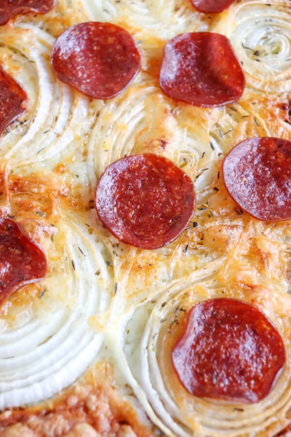 Close-up of a pepperoni pizza with visible mozzarella cheese, crispy onion ring chips, and herbs on a golden-brown crust. The pepperoni slices are arranged evenly across the surface.