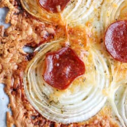 Close-up of a low-carb pizza crust topped with slices of pepperoni and onion rings. The crust appears crispy with melted cheese and browned edges. The pizza is placed on a sheet of baking parchment.