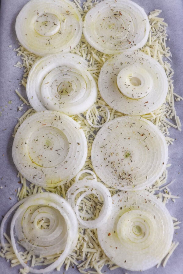 Slices of white onion arranged on a baking sheet lined with shredded cheese and sprinkled with herbs.