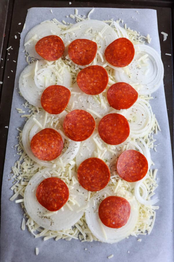 Unbaked pizza on parchment paper, topped with sliced onions, grated cheese, and scattered pepperoni slices.