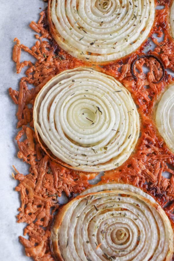 Close-up of baked onion slices with herbs on melted cheese, creating a crispy, browned edge. The cheese appears slightly golden, and the layers of the onion are distinct and well-cooked, placed on a parchment-lined surface.