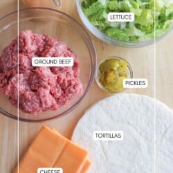 Ingredients for a dish displayed on a table: ground beef in a bowl, lettuce in another bowl, slices of cheddar cheese, tortillas, sliced pickles, and a small bowl labeled Big Mac Sauce.