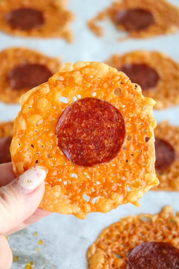 A hand holds a round, crispy cheese chip with a slice of pepperoni in the center. More similar chips are visible on a parchment-lined baking sheet in the background.