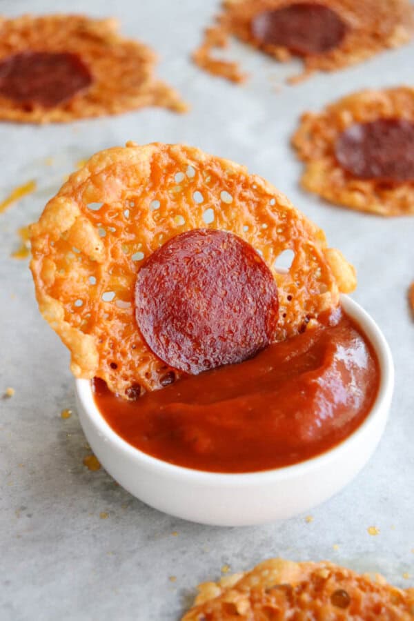 A round, crispy cheese and pepperoni chip is dipped into a small bowl of red marinara sauce, with more chips visible on a sheet of parchment paper in the background.
