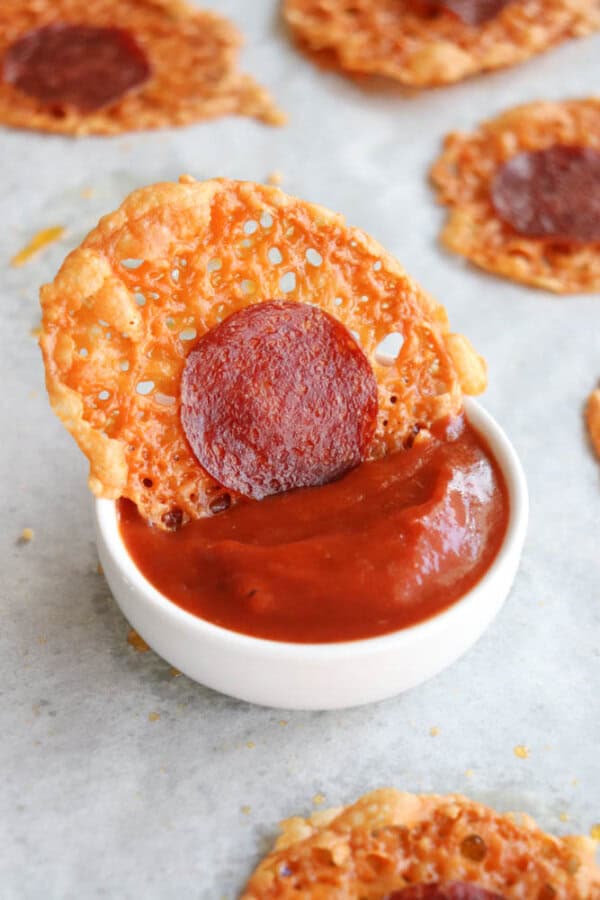 A crispy baked cheese round with a slice of pepperoni is being dipped into a small white bowl of red marinara sauce. More cheese rounds with pepperoni are in the background on parchment paper.
