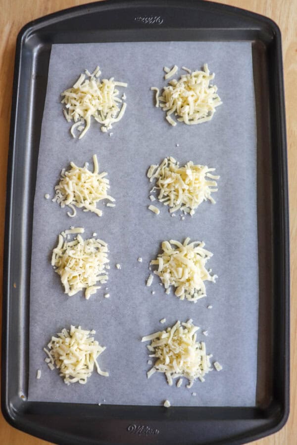A baking tray lined with parchment paper holds eight small piles of shredded cheese, arranged in two columns. The tray is on a wooden surface.