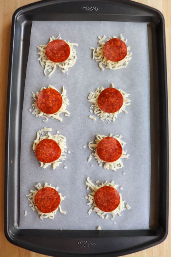 A baking tray lined with parchment paper holds eight small piles of shredded cheese, each topped with a single slice of pepperoni, spaced evenly in two rows.