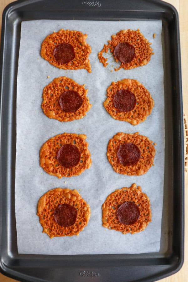 A baking tray lined with parchment paper holds eight round, lacy cheese crisps, each with a slice of pepperoni in the center. The crisps are unevenly shaped and spaced out on the tray.