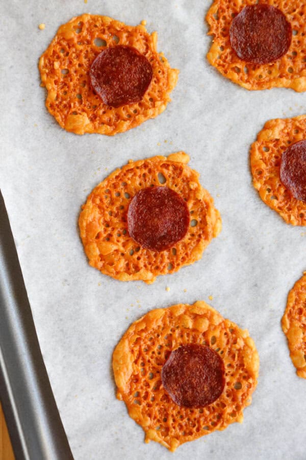 Five round, crispy cheese and pepperoni chips are arranged on a baking tray lined with parchment paper. The chips have a lacy, golden-brown texture with a slice of pepperoni in the center of each.