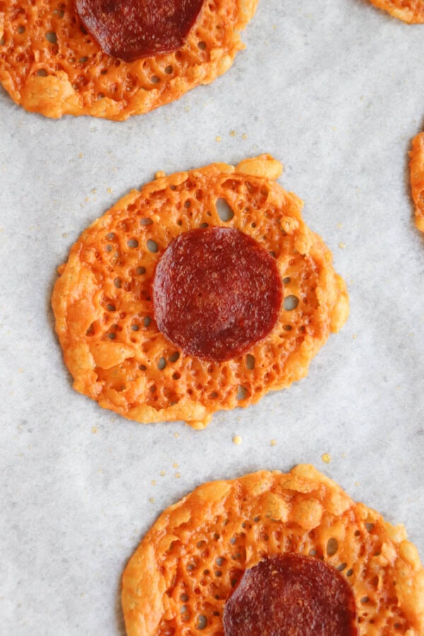 Close-up of round, lacy baked cheese crisps each topped with a slice of pepperoni, arranged on parchment paper.