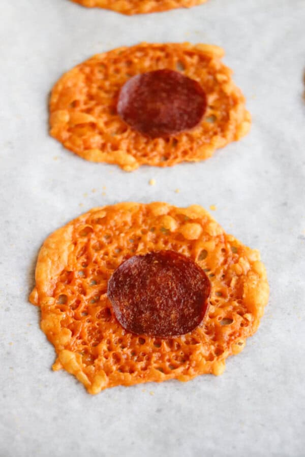Two round, crispy cheese chips topped with a single slice of pepperoni each, resting on a sheet of white parchment paper.
