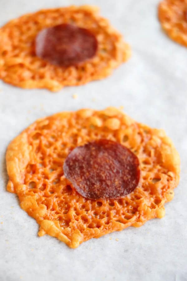 A close-up of a baked cheese crisp with a slice of pepperoni in the center, resting on a sheet of parchment paper. Another similar crisp is visible in the background.