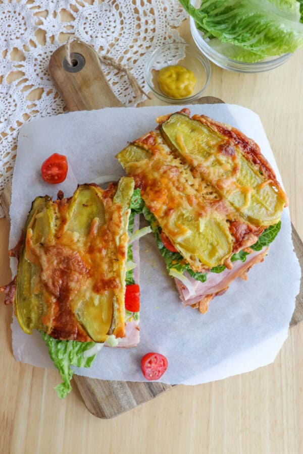 Two halves of a sandwich with melted cheese and pickles on top, filled with lettuce, ham, and tomato, are placed on parchment paper on a wooden board. A small bowl of mustard and loose lettuce are nearby.