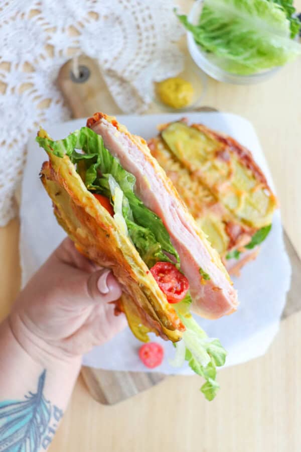 A hand holds a sandwich with layers of ham, lettuce, tomato, and pickles, using cheese crisps as bread. A bowl of lettuce and some mustard are in the background on a wooden board and lace tablecloth.