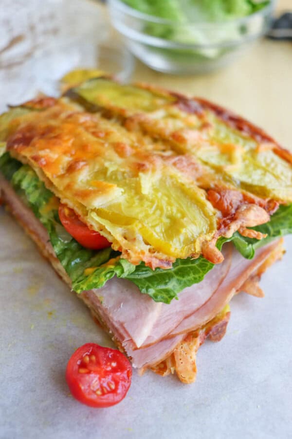 A close-up of a sandwich with crispy melted cheese as the bread, filled with lettuce, tomato, and thick slices of ham, placed on parchment paper with a tomato slice in front.