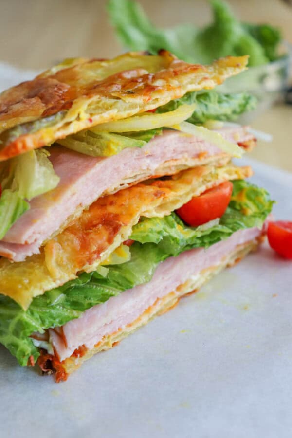 A sandwich made with crispy cheese slices as the bread, filled with layers of ham, lettuce, and tomato, sits on parchment paper with loose lettuce in the background.