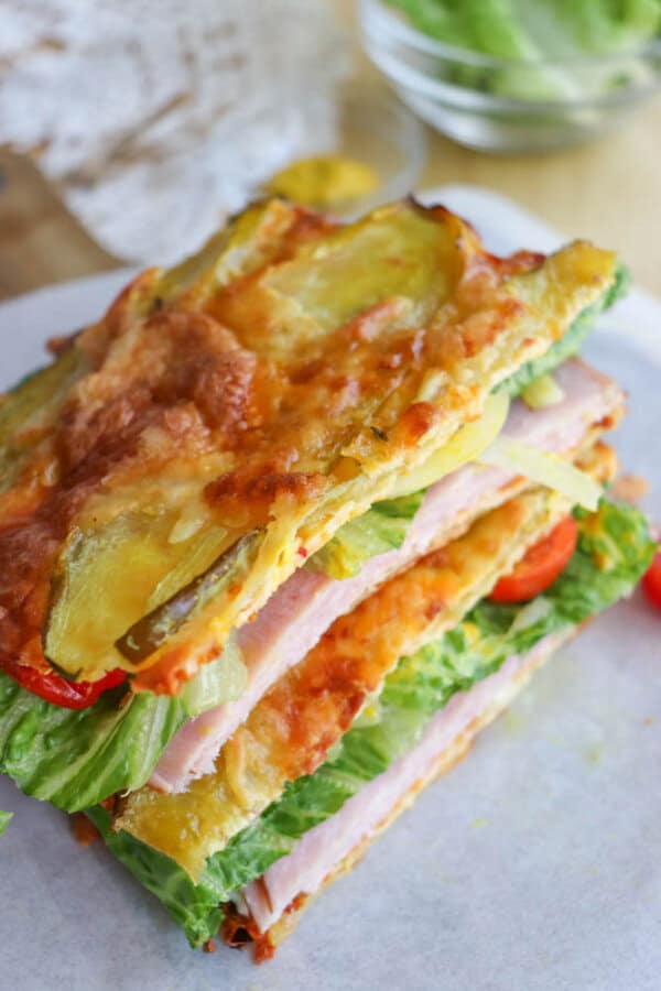 A stack of sandwiches made with layers of baked cheese and zucchini instead of bread, filled with ham, lettuce, and tomato, placed on parchment paper with a bowl of greens in the background.