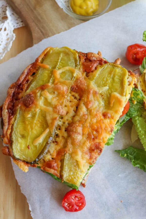 A slice of cheesy, baked casserole topped with pickle slices, melted cheddar cheese, and served with lettuce and cherry tomato halves on a piece of parchment paper. A small dish of mustard is in the background.