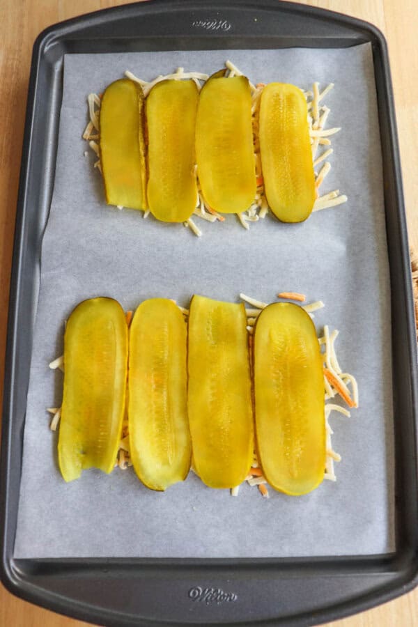 A baking tray lined with parchment paper holds two piles of shredded cheese topped with sliced dill pickles, ready to bake.