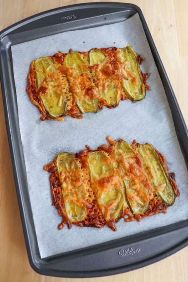 Two rectangular cheesy baked snacks topped with sliced green pickles are on a parchment-lined baking sheet. The snacks are golden brown with melted cheese bubbling around the pickles.