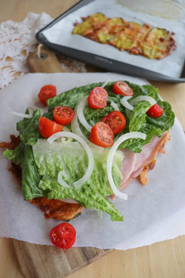 An open-faced sandwich on parchment paper topped with lettuce, sliced cherry tomatoes, onions, ham, and cheese, with a tray of baked food in the background on a wooden table.