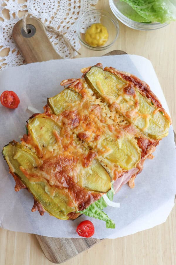 A baked sandwich topped with melted cheese and sliced pickles sits on parchment paper, garnished with lettuce and cherry tomatoes. A small bowl of mustard and lettuce leaves are nearby on a wooden table.