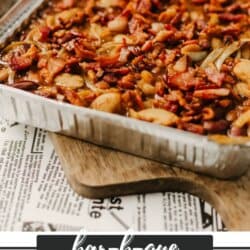 A foil tray filled with barbecued baked beans, topped with bacon, sits on a wooden board. Text on the image reads bar-b-que BAKED BEANS and homemadeheather.com.