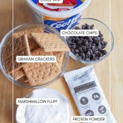 A flat lay of ingredients for a dessert recipe: a tub of Cool Whip, a bowl of chocolate chips, graham crackers, marshmallow fluff in a jar, and a packet of chocolate protein powder.