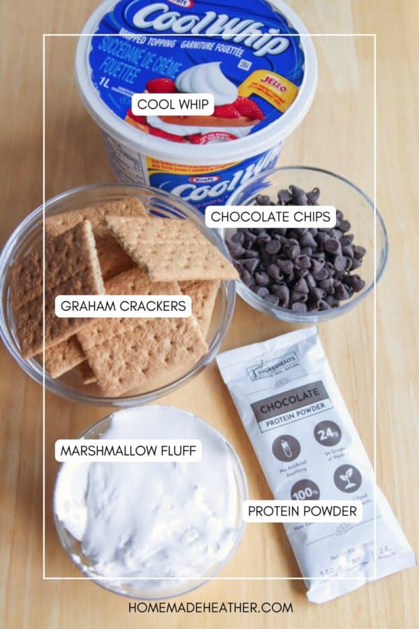 A flat lay of ingredients for a dessert recipe: a tub of Cool Whip, a bowl of chocolate chips, graham crackers, marshmallow fluff in a jar, and a packet of chocolate protein powder.