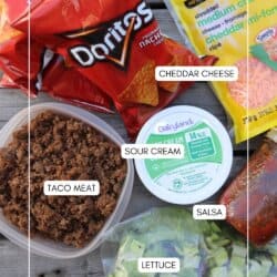 An overhead view of ingredients for taco salad and Walking Tacos Recipe: bags of Doritos chips, shredded cheddar cheese, cooked taco meat, sour cream, salsa, and chopped lettuce, all arranged on a wooden surface.