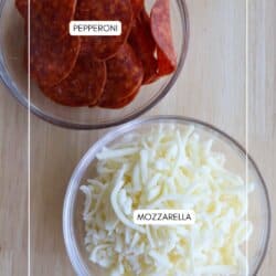 Two glass bowls on a wooden surface: one filled with sliced pepperoni, and the other with shredded mozzarella cheese. Each bowl is labeled with its contents.