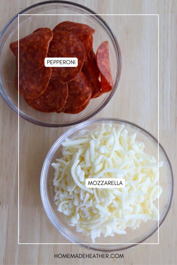Two glass bowls on a wooden surface: one filled with sliced pepperoni, and the other with shredded mozzarella cheese. Each bowl is labeled with its contents.
