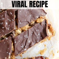 Close-up of homemade Snickers date bars on parchment paper. The bars are topped with a layer of chocolate and sprinkled with coarse salt, with a textured surface that reveals chunks of nuts and caramel. Overlay text reads, Snickers Date Bars Viral Recipe.