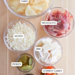 A wooden table displays bowls labeled with various foods: chips, prosciutto, mozzarella, brie, pickles, cowboy candy, and jam. Each item is in a clear glass bowl.
