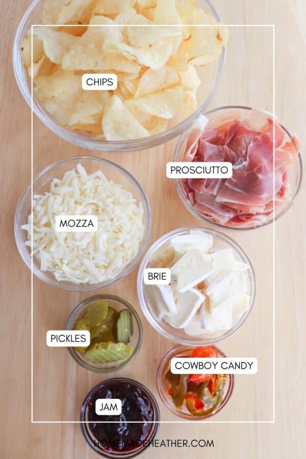A wooden table displays bowls labeled with various foods: chips, prosciutto, mozzarella, brie, pickles, cowboy candy, and jam. Each item is in a clear glass bowl.