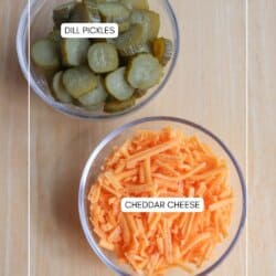 Two clear bowls on a wooden surface: one contains sliced dill pickles and the other contains shredded cheddar cheese. Labels identify each bowls contents.