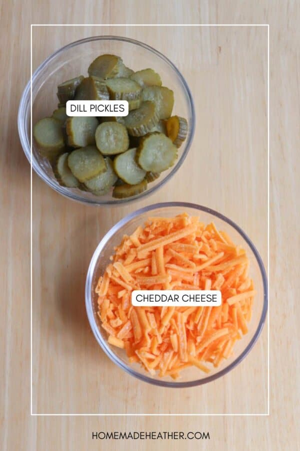 Two clear bowls on a wooden surface: one contains sliced dill pickles and the other contains shredded cheddar cheese. Labels identify each bowls contents.