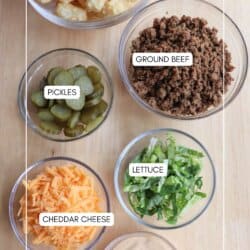 Bowls of ingredients on a wooden surface: tater tots, ground beef, pickles, shredded cheddar cheese, chopped lettuce, and sauce. Each bowl is labeled accordingly.
