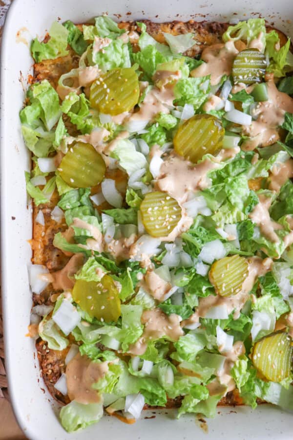 A casserole dish filled with a layered salad, topped with chopped lettuce, sliced pickles, diced onions, and a drizzle of creamy dressing.
