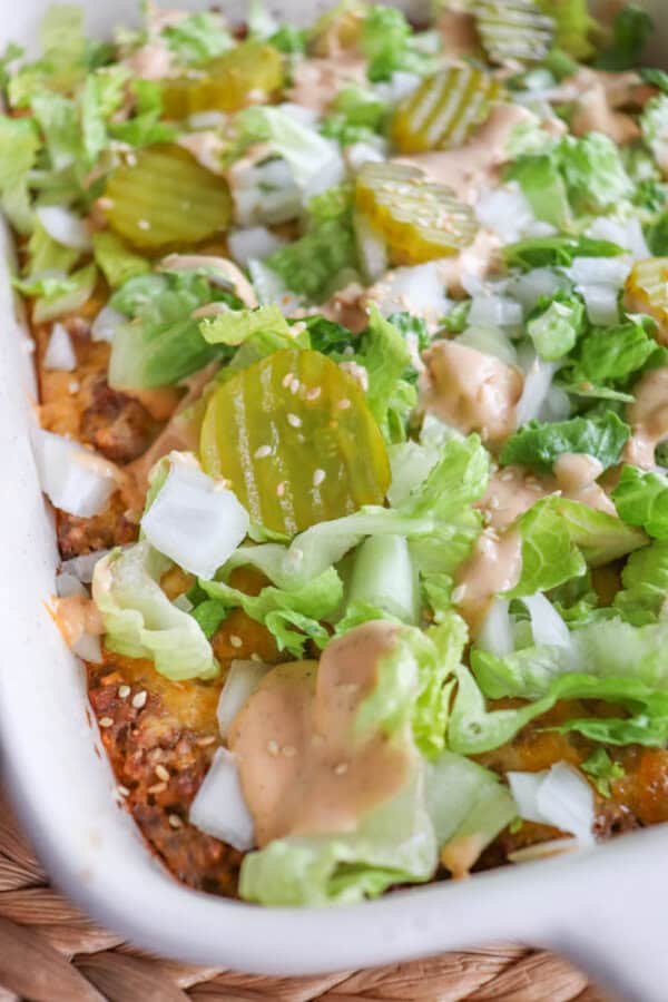 A close-up of a dish topped with lettuce, sliced pickles, chopped onions, and a creamy sauce, resembling a cheeseburger casserole. The dish is in a white baking dish, showcasing a mix of textures and vibrant colors.