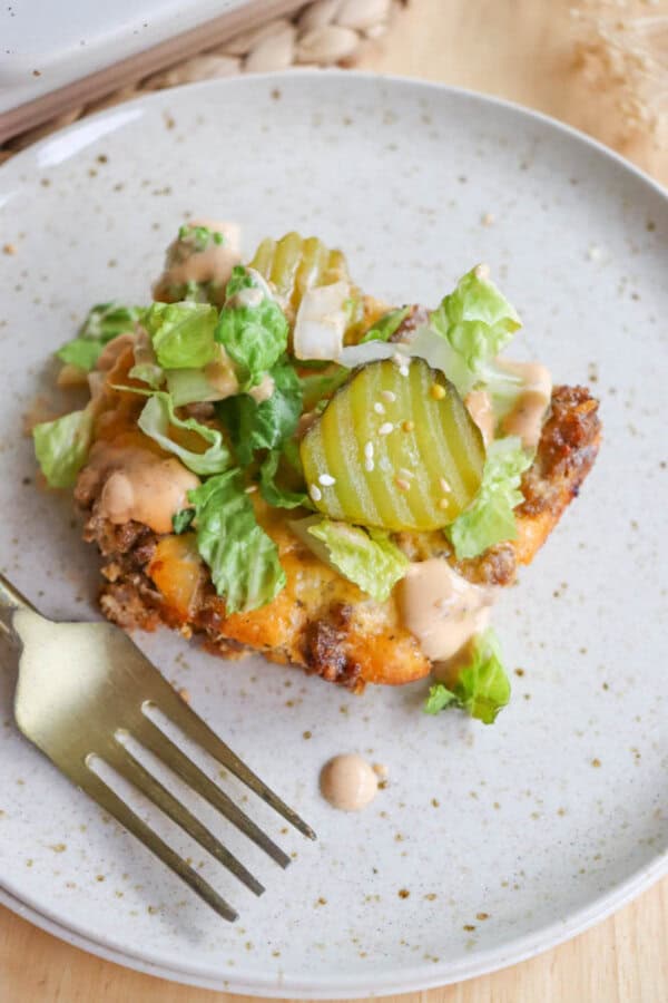 A plate with a slice of cheeseburger casserole topped with shredded lettuce, sliced pickles, and drizzled sauce. A fork is placed beside the casserole on a speckled ceramic plate.