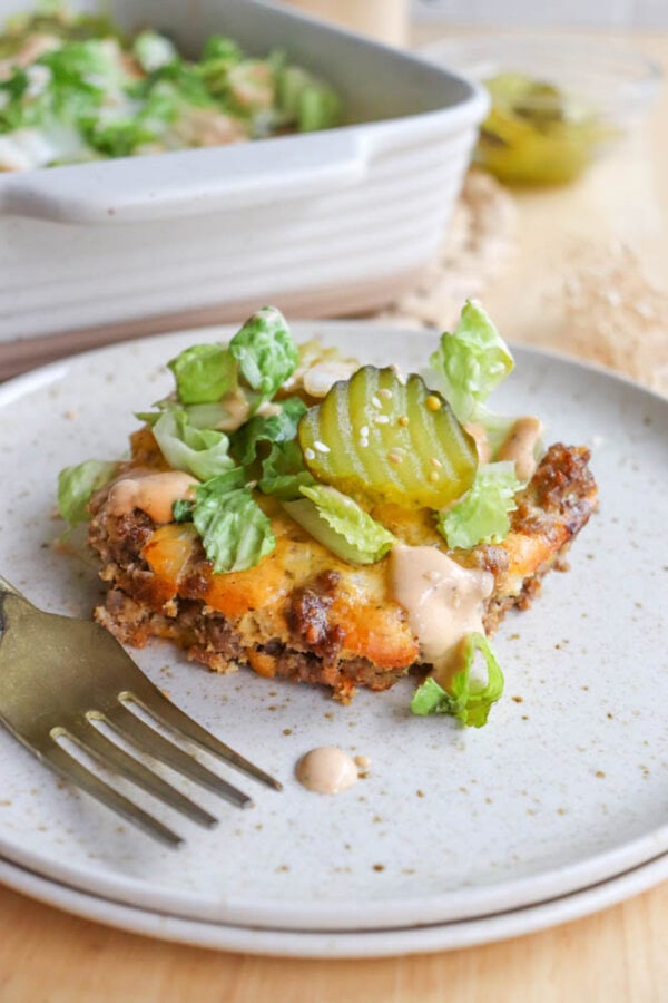 A slice of casserole served on a speckled plate, topped with melted cheese, chopped lettuce, pickles, and sauce. A gold fork rests beside it. A dish with more casserole is blurred in the background.