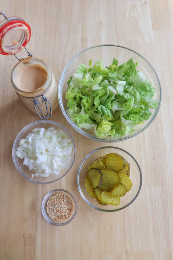 A top-down view of ingredients on a wooden surface: a jar of sauce, a bowl of chopped lettuce, diced onions in a bowl, a bowl of pickle slices, and a small dish of sesame seeds.