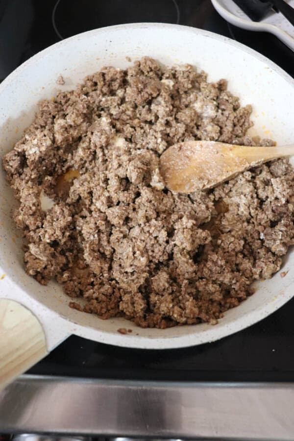 A white skillet on a stovetop holds browned ground meat, being stirred with a wooden spoon. The meat appears seasoned and cooked, with a slightly crumbly texture.