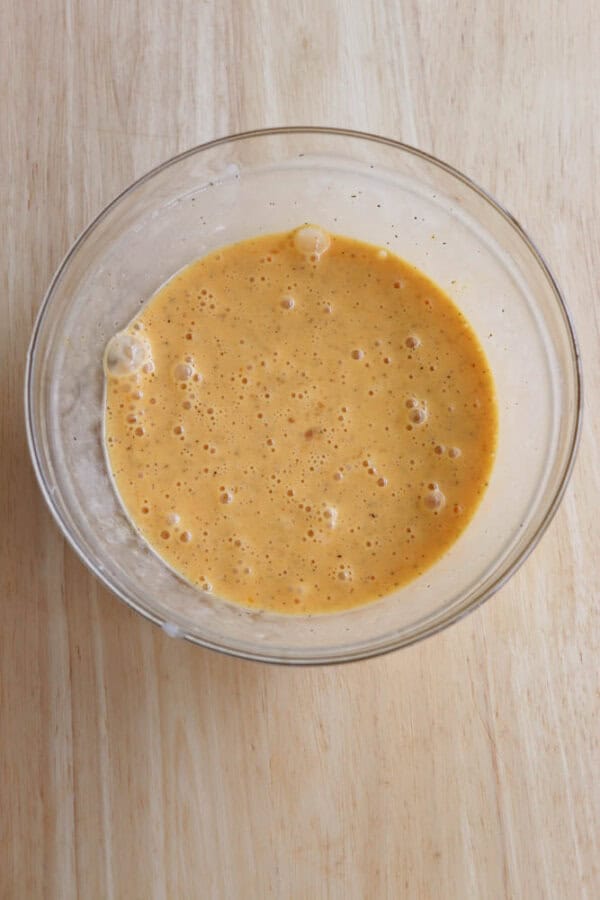 A glass bowl filled with a frothy, orange batter mixture sits on a light wooden surface. Small bubbles are visible on the batters surface, indicating it is well-mixed and airy.