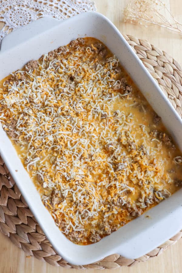 A baked casserole dish filled with noodles, ground meat, and topped with melted cheese. The dish is set on a woven placemat with a lace doily partially visible in the background.