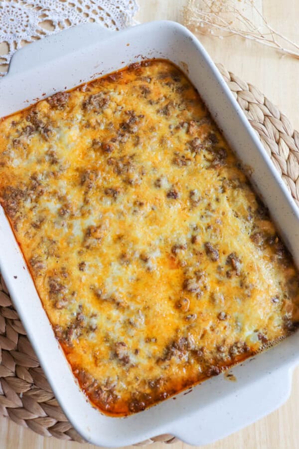 Casserole dish filled with baked shepherds pie, topped with melted cheese and browned bits. The dish rests on a woven placemat with a lace cloth partially visible in the upper left corner.
